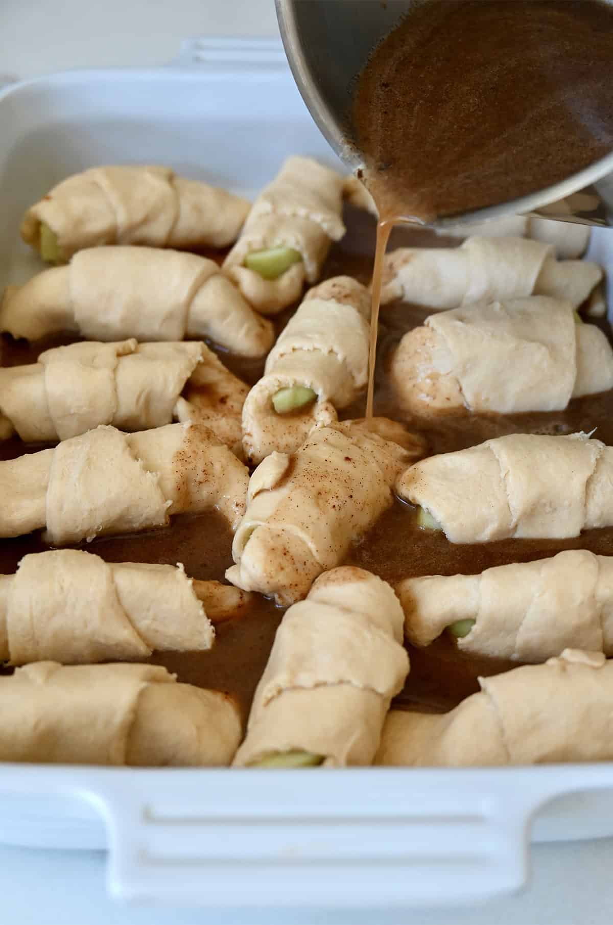 Brown sugar and butter sauce being poured over crescent roll apple dumplings before baking.