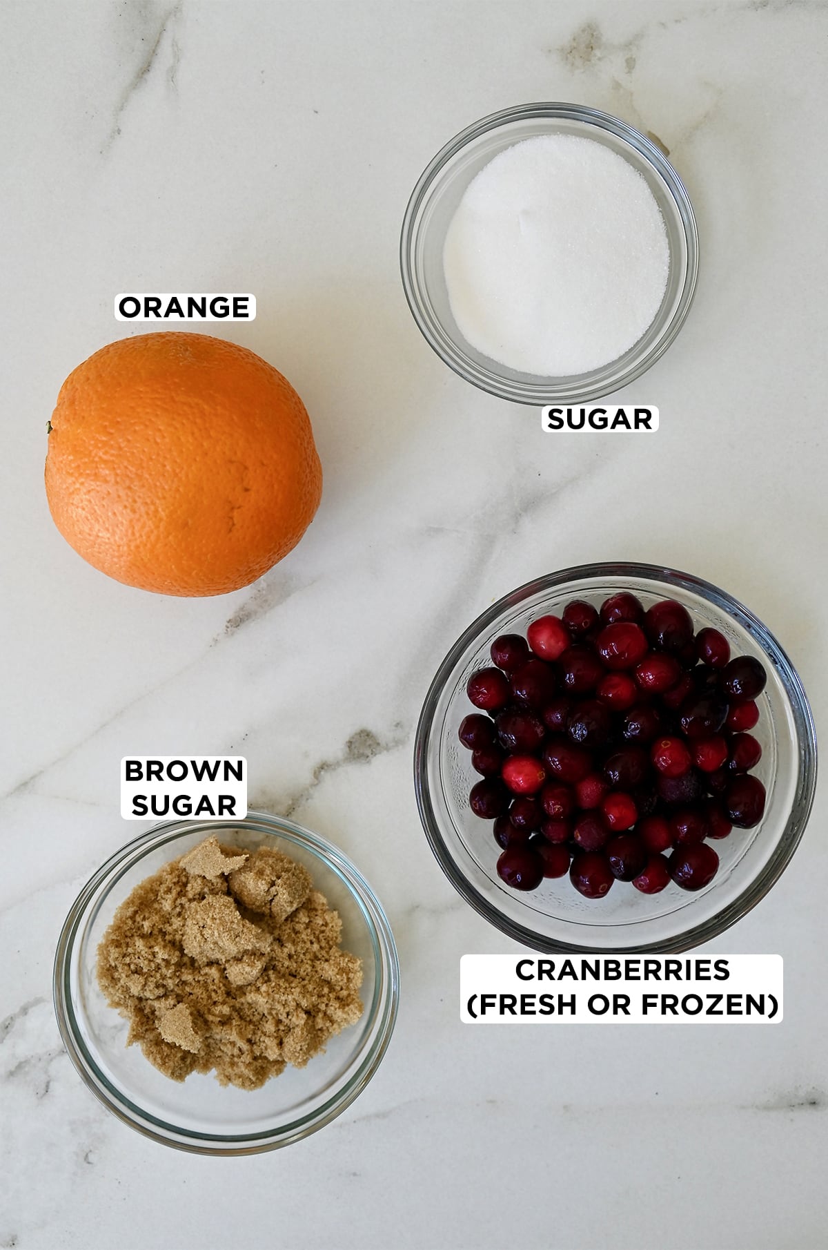 An orange next to three bowls containing white sugar, fresh cranberries and brown sugar.