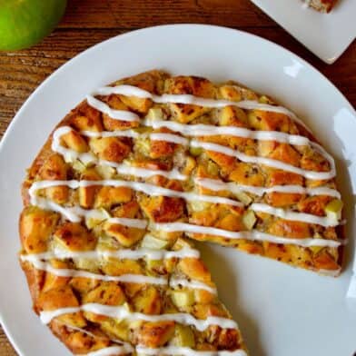 An icing-drizzled Apple Cinnamon Roll Bake is on a white plate, with a wedge missing. In the background are a couple of Granny Smith apples.