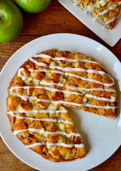 An icing-drizzled Apple Cinnamon Roll Bake is on a white plate, with a wedge missing. In the background are a couple of Granny Smith apples.