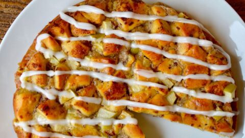An icing-drizzled Apple Cinnamon Roll Bake is on a white plate, with a wedge missing. In the background are a couple of Granny Smith apples.