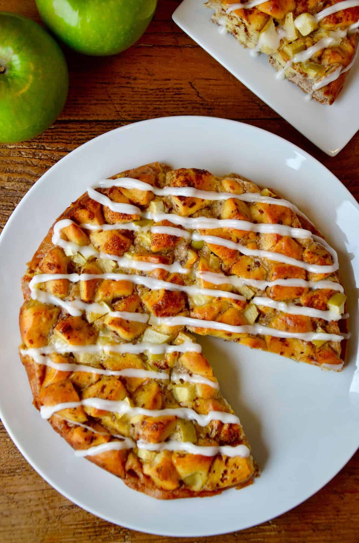 An icing-drizzled Apple Cinnamon Roll Bake is on a white plate, with a wedge missing. In the background are a couple of Granny Smith apples.