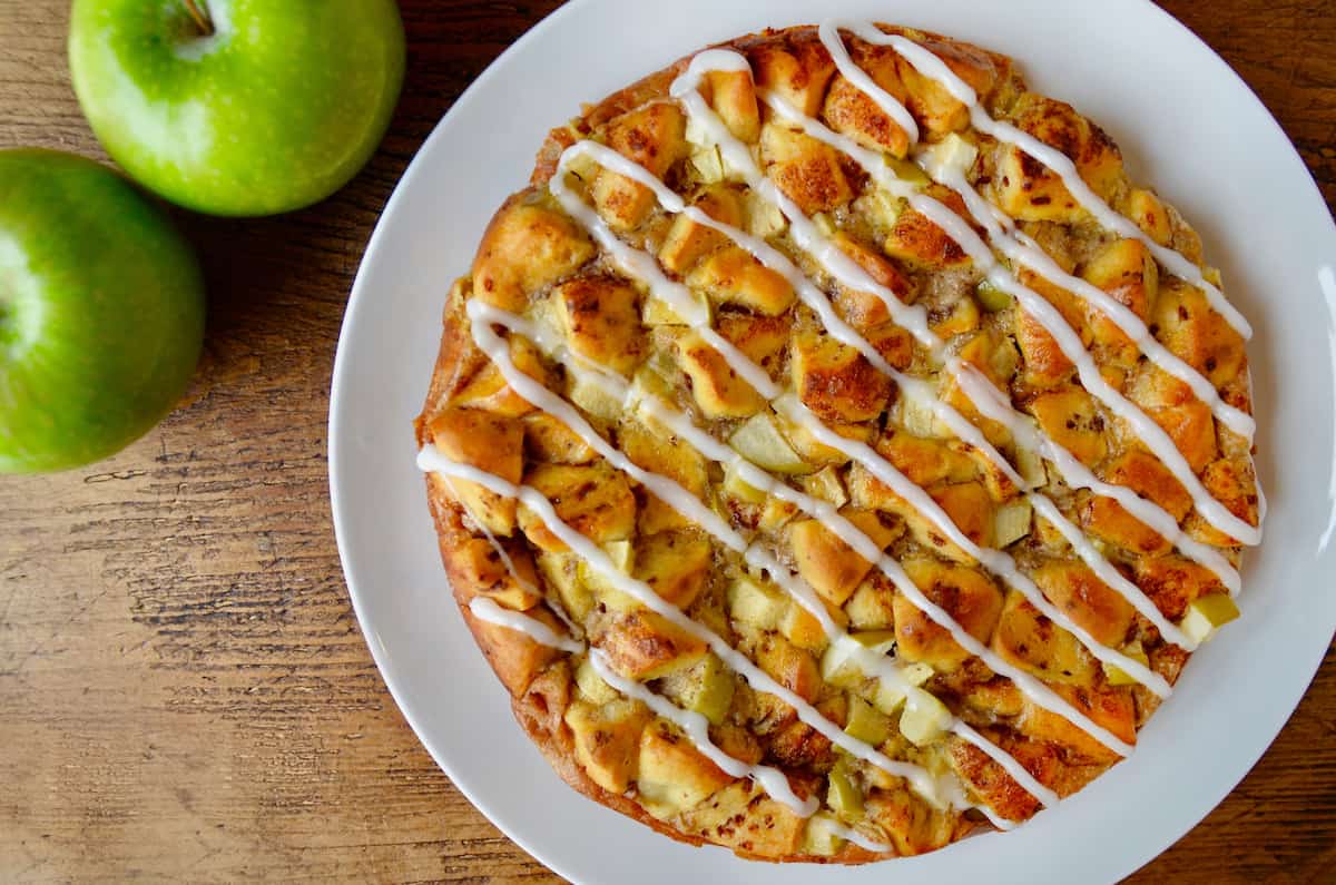 An icing-drizzled Apple Cinnamon Roll Bake is on a white plate, with a couple of Granny Smith apples in the background.