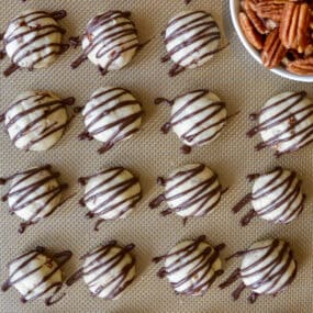 Rows of chocolate-drizzled pecan cookie balls are on a Silpat-lined baking sheet. A bowl of pecan halves sits nearby.
