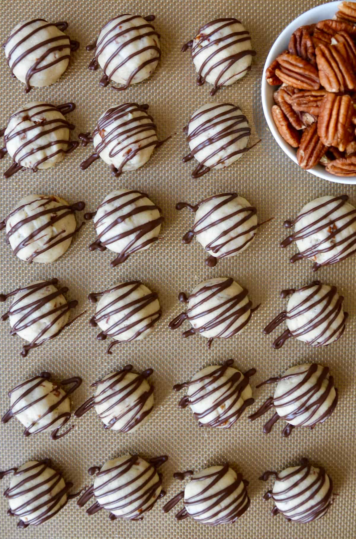 Rows of chocolate-drizzled pecan cookie balls are on a Silpat-lined baking sheet. A bowl of pecan halves sits nearby.