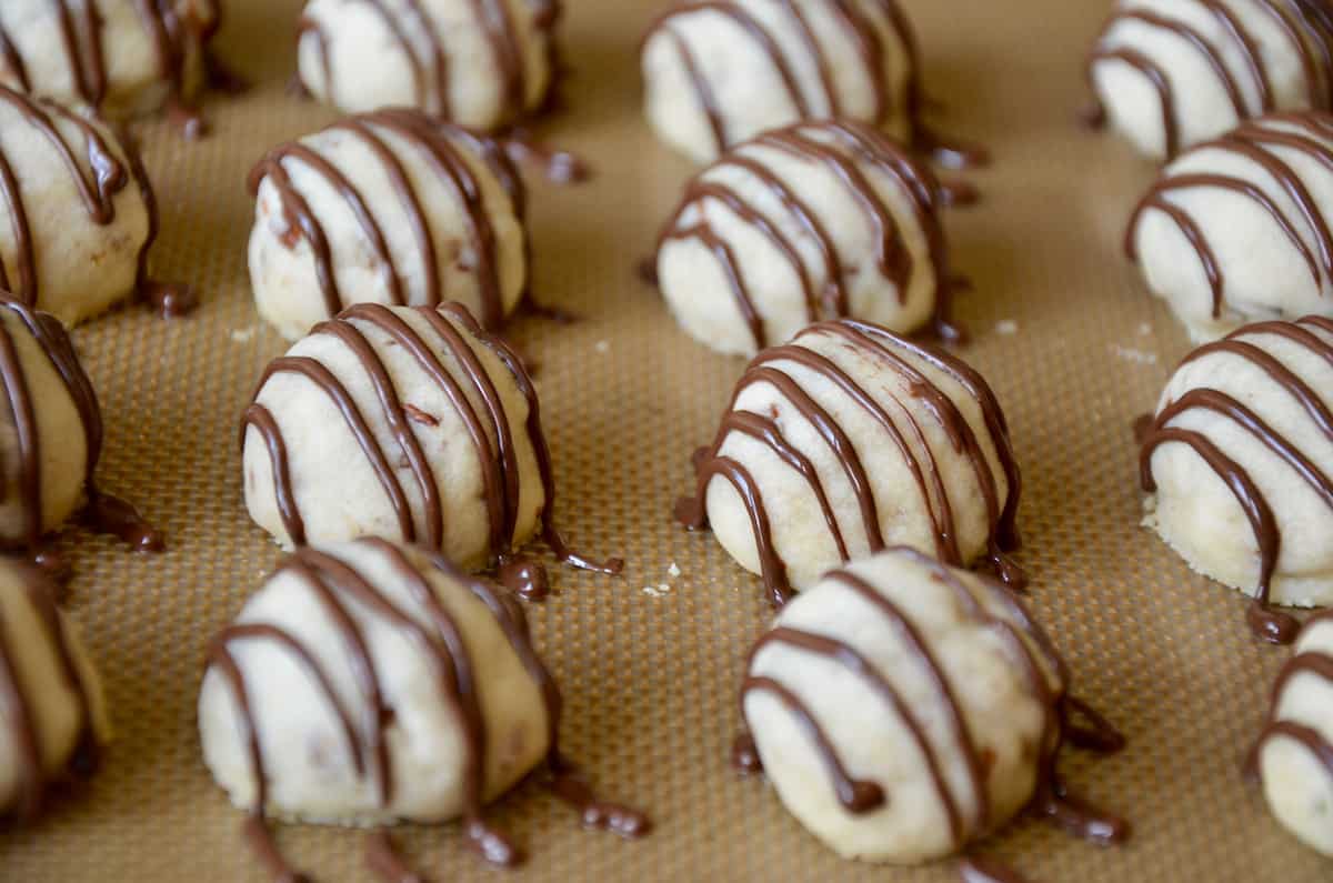 Rows of chocolate-drizzled pecan cookie balls on a Silpat-lined baking sheet. 