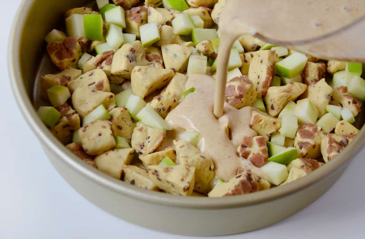 Custard is being poured over a mixture of chopped apples and cinnamon roll dough in a round cake pan.