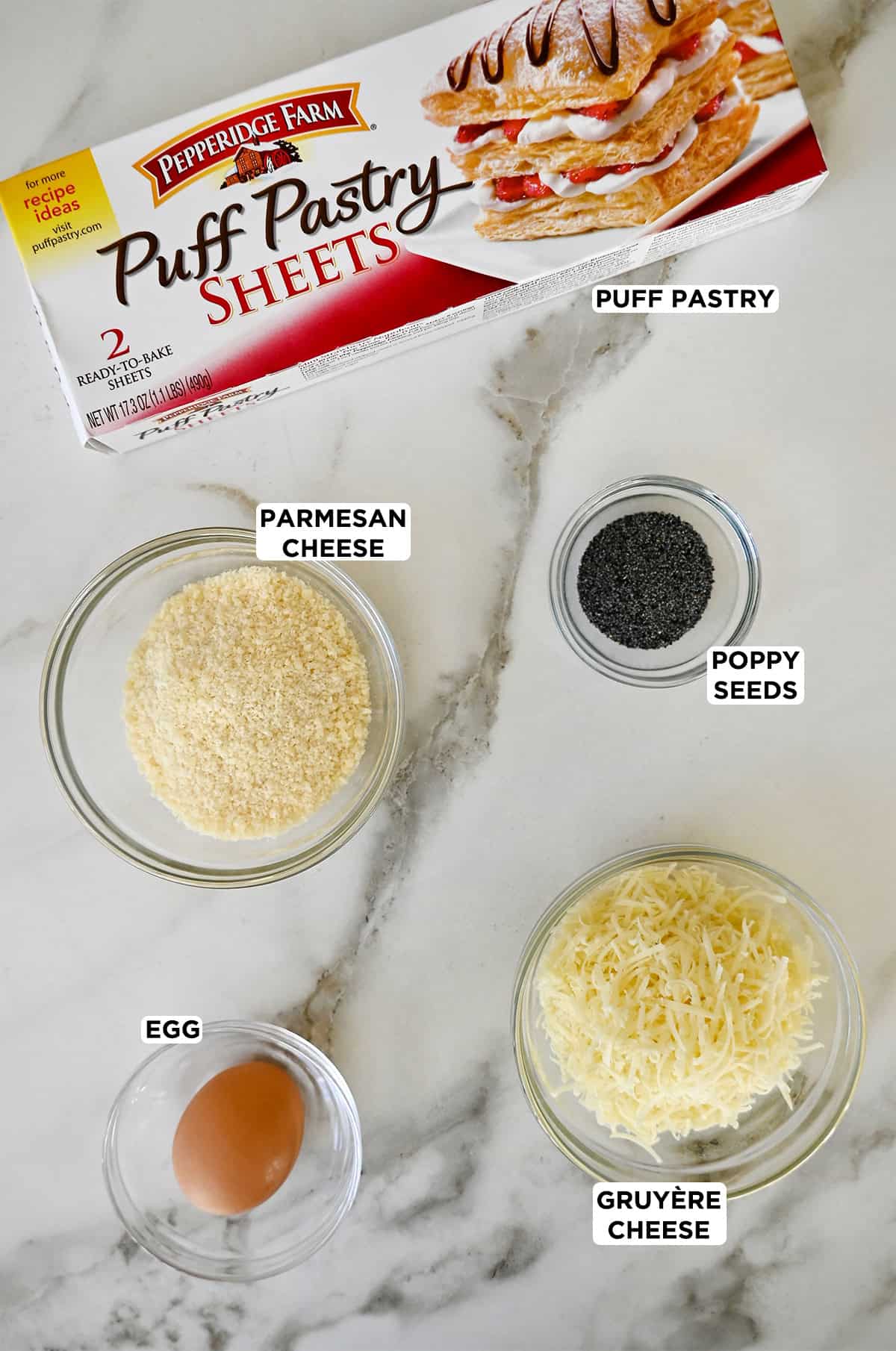 Ingredients to make cheese straws, including a box of frozen puff pastry, grated parmesan and Gruyère cheeses, poppy seeds, and an egg.