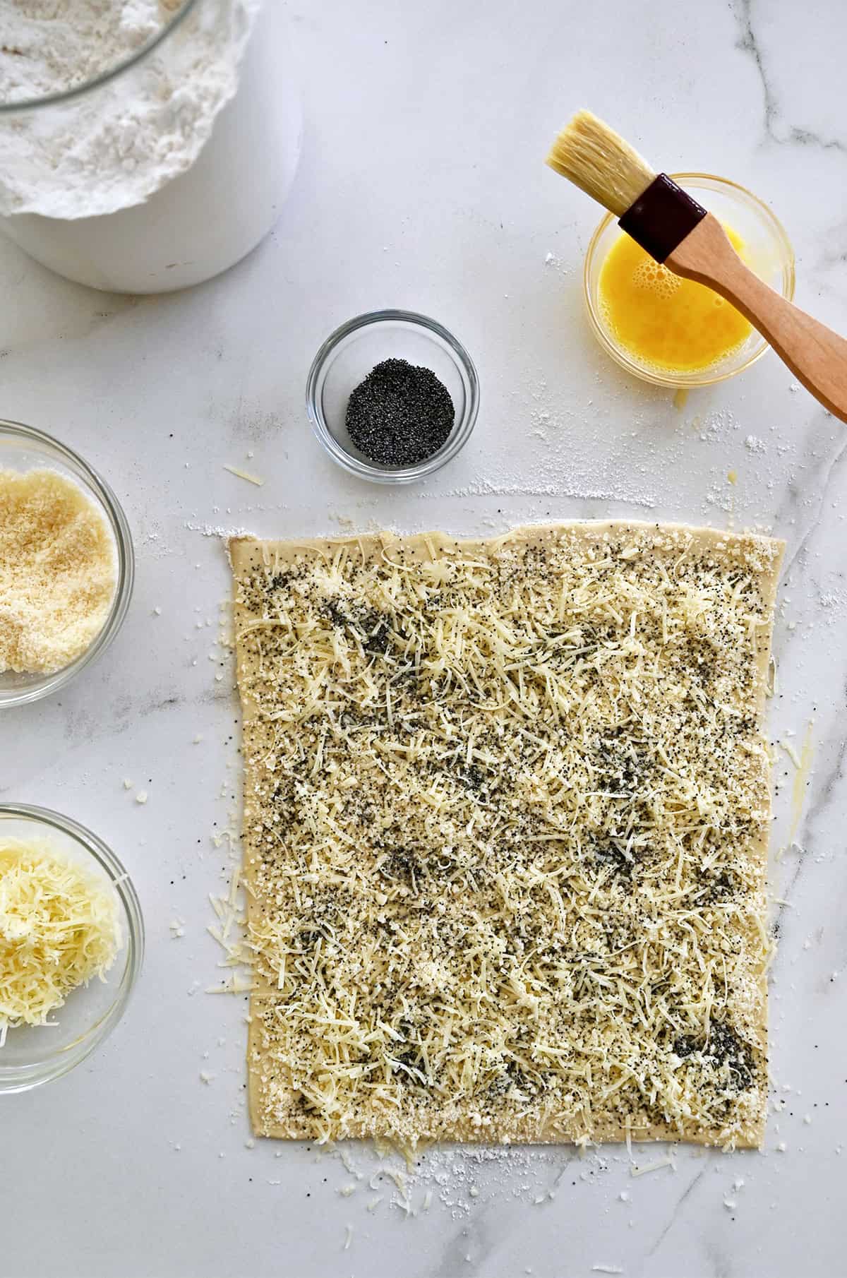 A sheet of puff pastry rolled out, topped with grated cheese and poppy seeds.