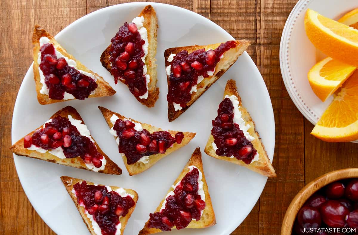 A small bowl filled with fresh cranberries is next to goat cheese toast topped with orange cranberry sauce and pomegranate arils on a white plate.