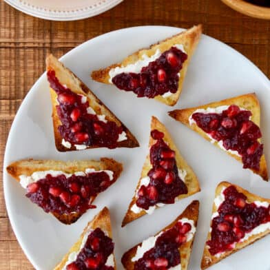 Orange cranberry sauce and pomegranate arils atop goat cheese toast on a white plate.