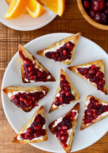Orange cranberry sauce and pomegranate arils atop goat cheese toast on a white plate.