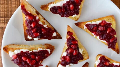 Orange cranberry sauce and pomegranate arils atop goat cheese toast on a white plate.