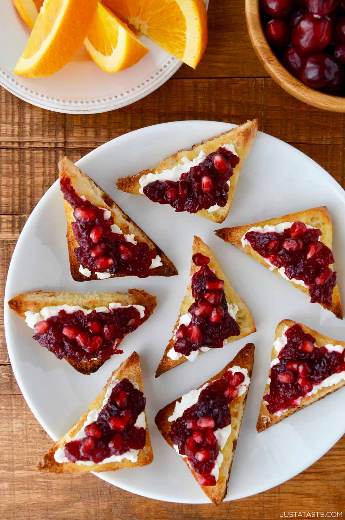 Orange cranberry sauce and pomegranate arils atop goat cheese toast on a white plate.