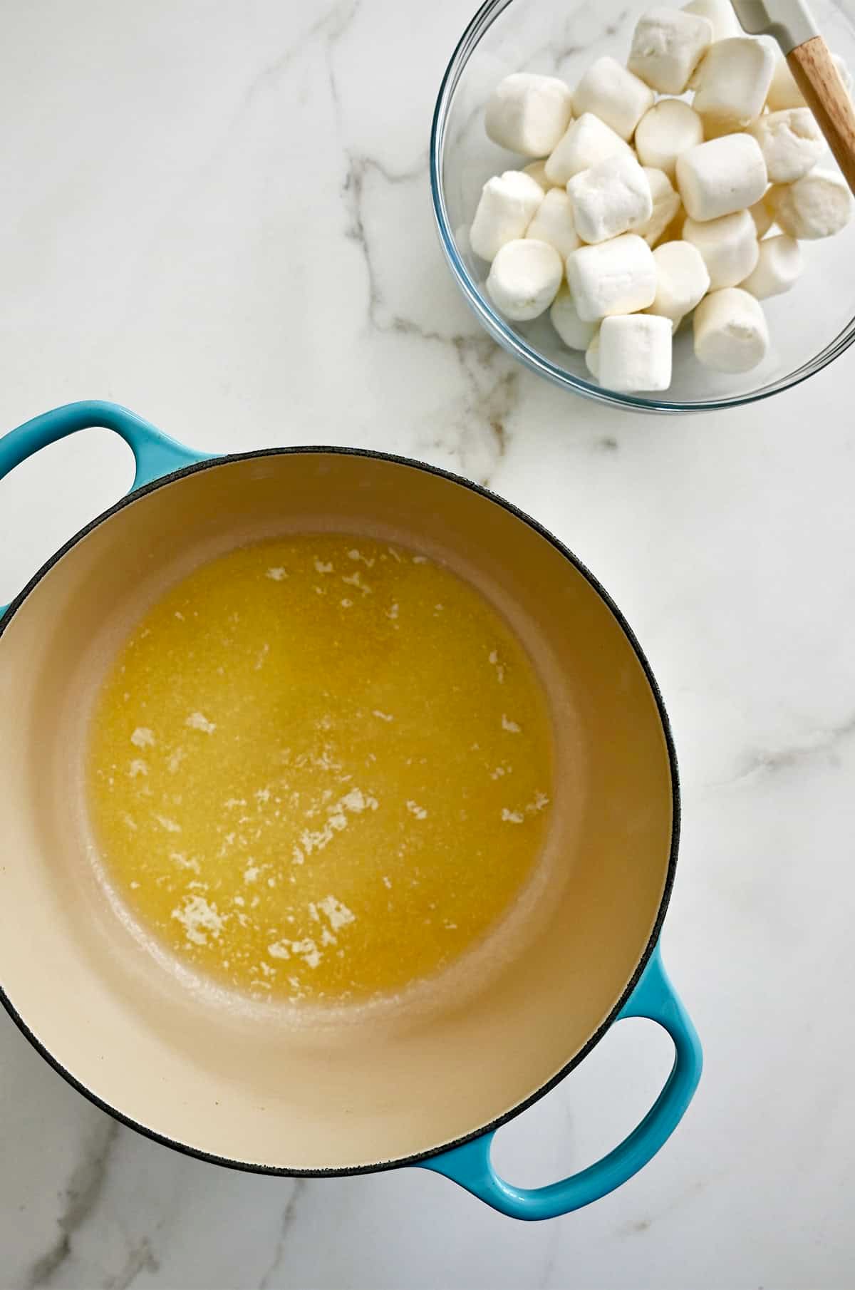Melted butter in a large pot next to a bowl containing large marshmallows.