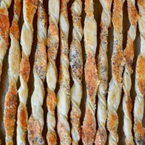 A close up of cheesy puff pastry twists on a baking sheet lined with parchment paper