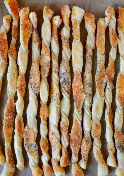A close up of cheesy puff pastry twists on a baking sheet lined with parchment paper