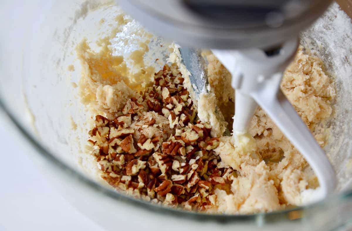 In the glass bowl of a stand mixer, chopped pecans are about to be mixed into cookie dough.