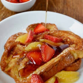 On a white plate, challah French toast is topped with sliced strawberries and orange segments and is being drizzled with maple syrup.