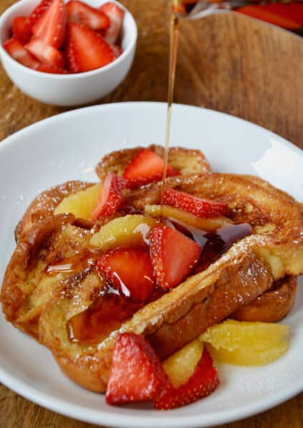 On a white plate, challah French toast is topped with sliced strawberries and orange segments and is being drizzled with maple syrup.