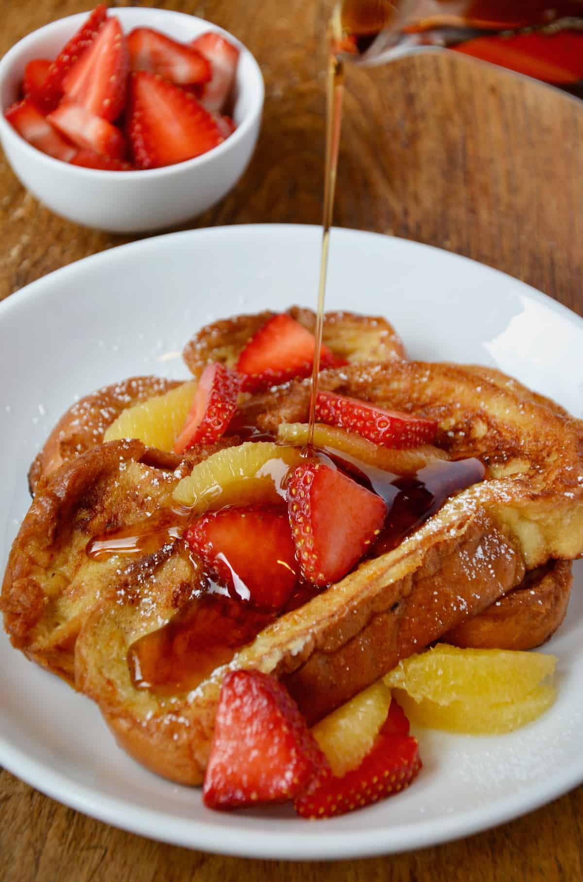On a white plate, challah French toast is topped with sliced strawberries and orange segments and is being drizzled with maple syrup.
