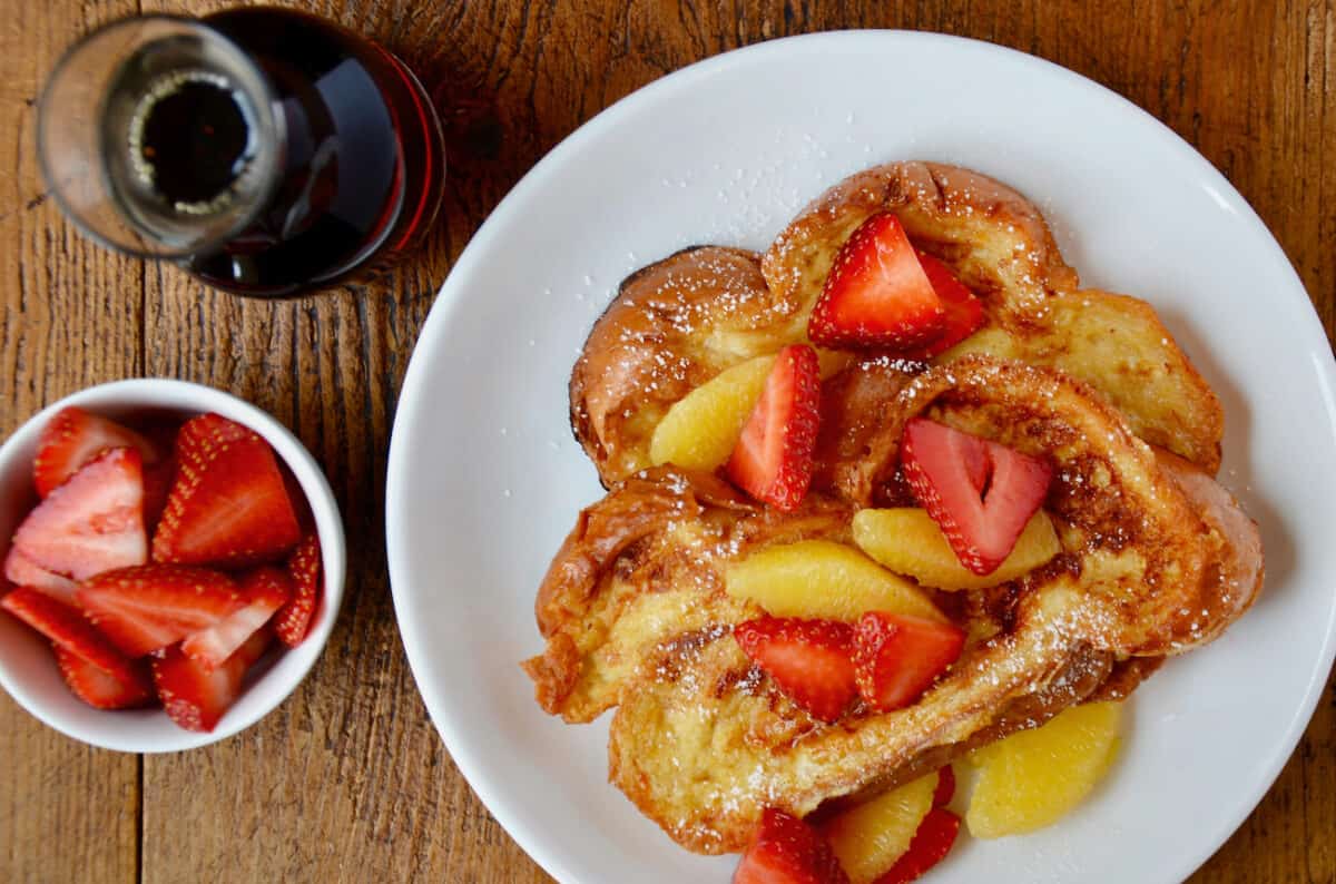 Challah French toast is topped with strawberries and orange segments. Nearby is a dish of strawberries and a glass pitcher of maple syrup.