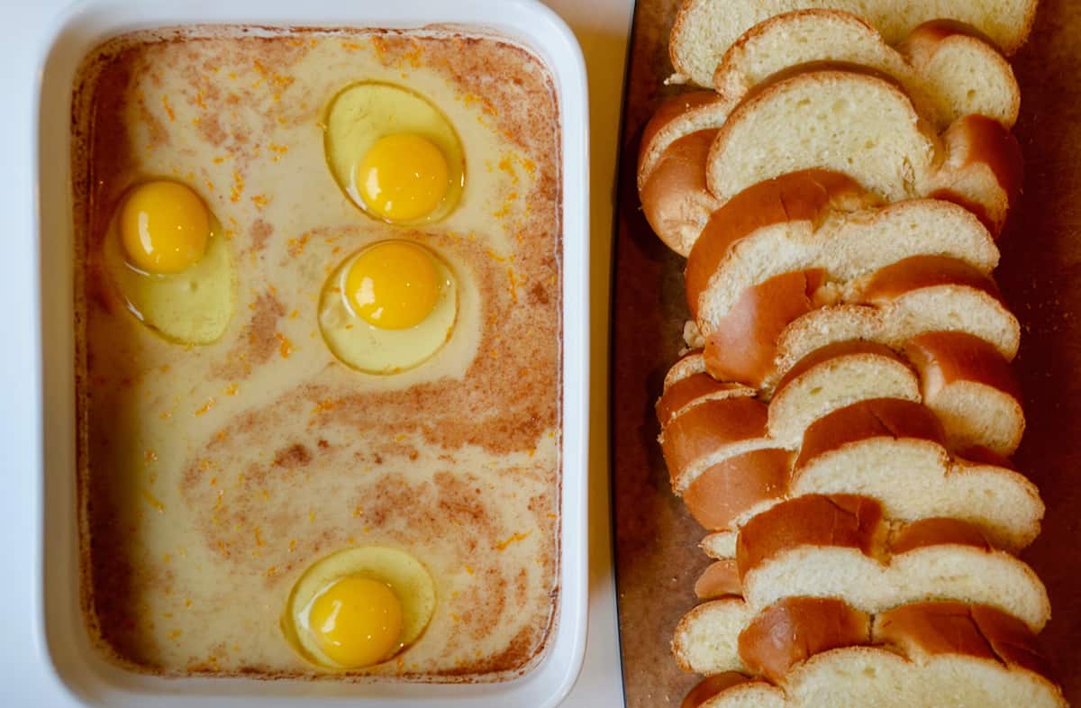 A shallow dish holds eggs, milk, cinnamon, brown sugar and other ingredients. Next to it is sliced challah bread.