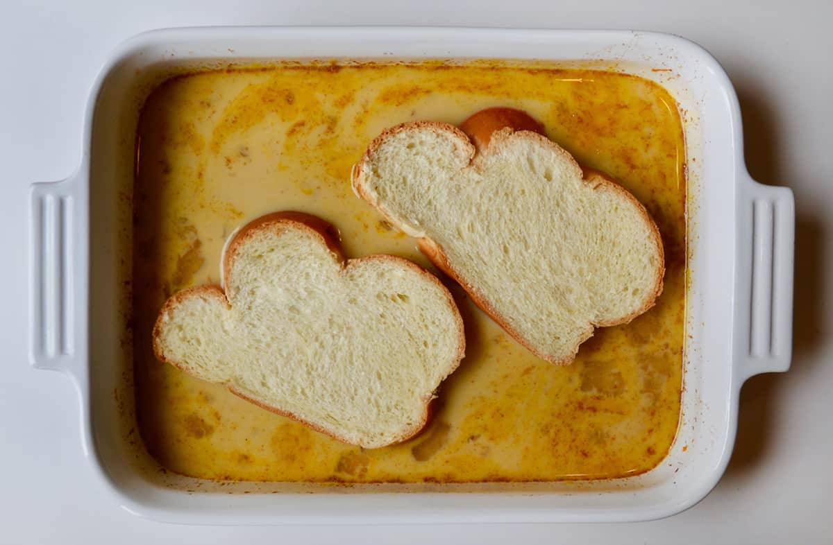 Two pieces of challah bread sit in a custard mixture in a white rectangular dish.