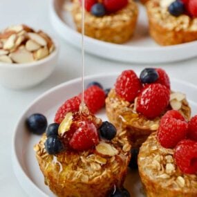 Baked Oatmeal Cups on a white plate topped with fresh raspberries and blueberries being drizzled with warm maple syrup