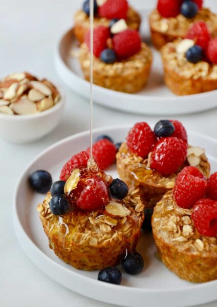 Baked Oatmeal Cups on a white plate topped with fresh raspberries and blueberries being drizzled with warm maple syrup