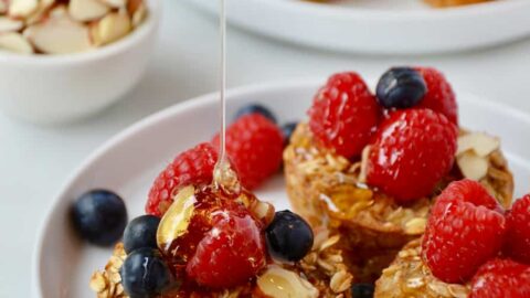 Baked Oatmeal Cups on a white plate topped with fresh raspberries and blueberries being drizzled with warm maple syrup