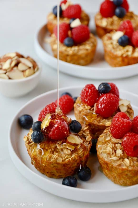 Baked Oatmeal Cups on a white plate topped with fresh raspberries and blueberries being drizzled with warm maple syrup