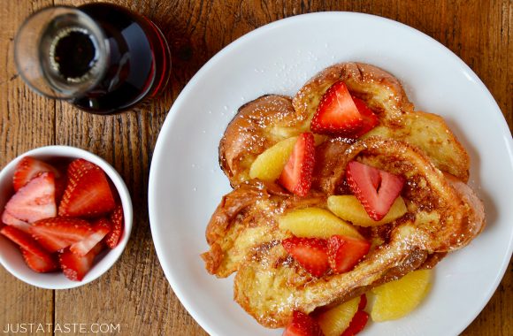 A white plate with Challah French Toast topped with fruit and a carafe of syrup