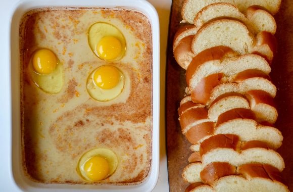 A white dish containing eggs and heavy cream alongside sliced Challah bread on a wood cutting board