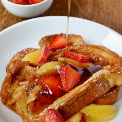 A white plate containing Challah French Toast with syrup being drizzled on top