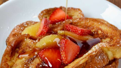 A white plate containing Challah French Toast with syrup being drizzled on top