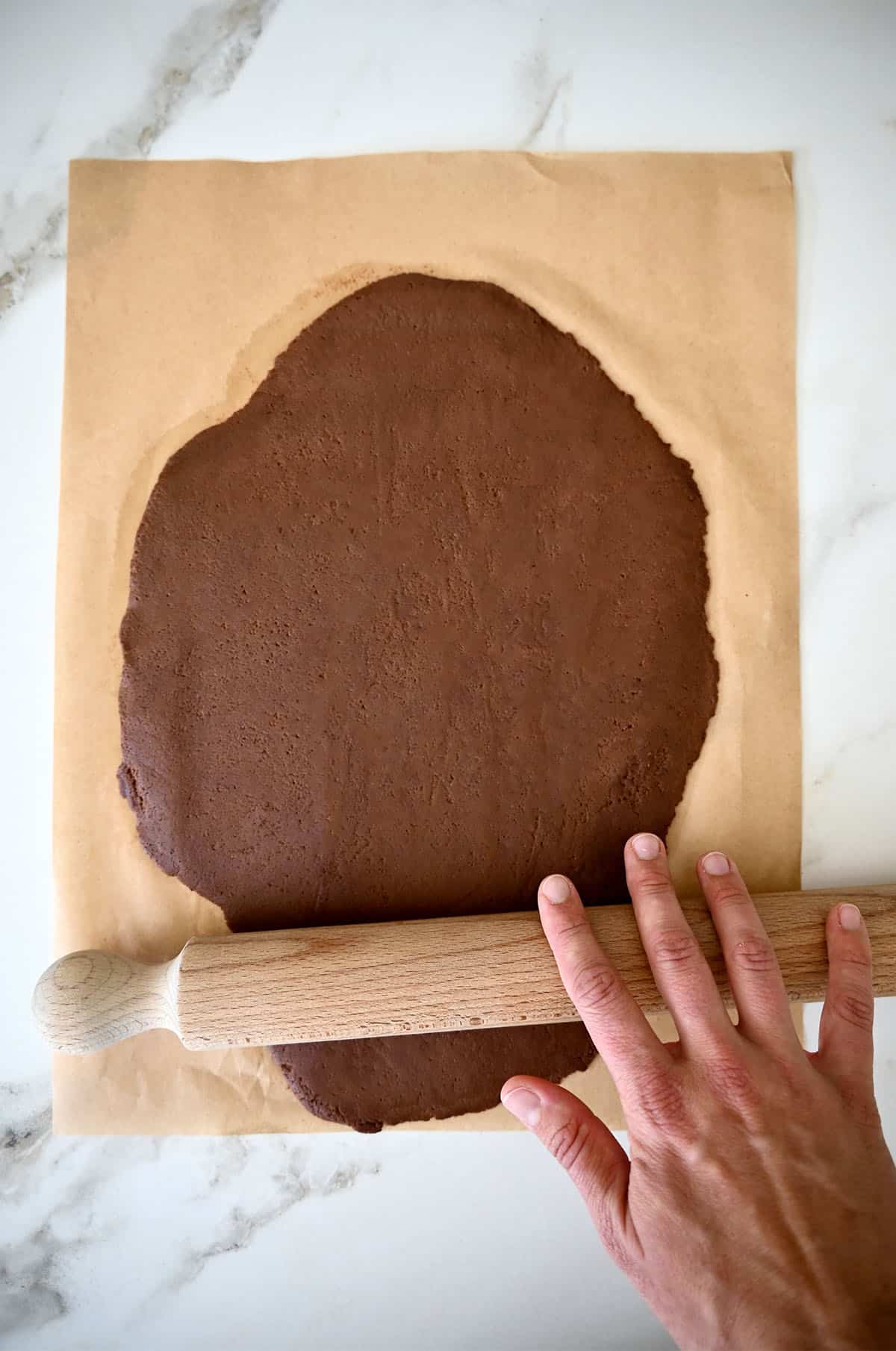 A hand holds a rolling pin atop chocolate shortbread dough on parchment paper.