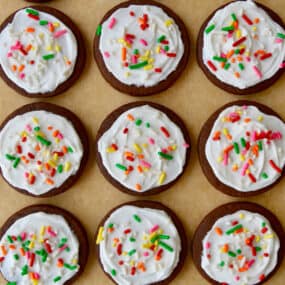 Rows of chocolate sugar cookies topped with vanilla buttercream frosting and rainbow sprinkles atop brown parchment paper.