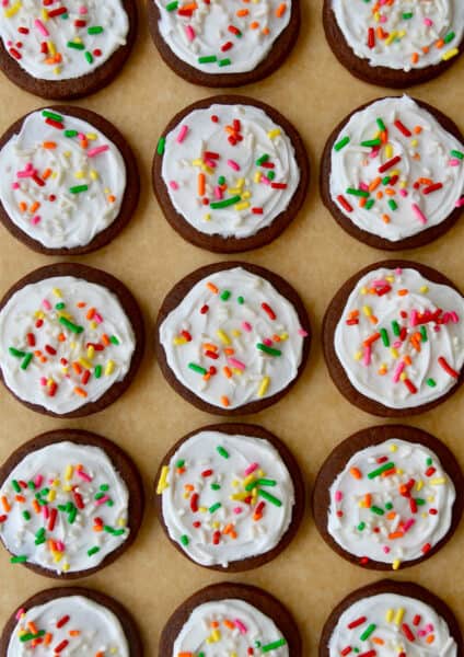 Rows of chocolate sugar cookies topped with vanilla buttercream frosting and rainbow sprinkles atop brown parchment paper.
