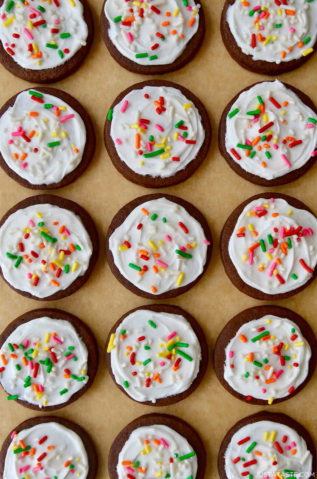 Rows of chocolate sugar cookies topped with vanilla buttercream frosting and rainbow sprinkles atop brown parchment paper.