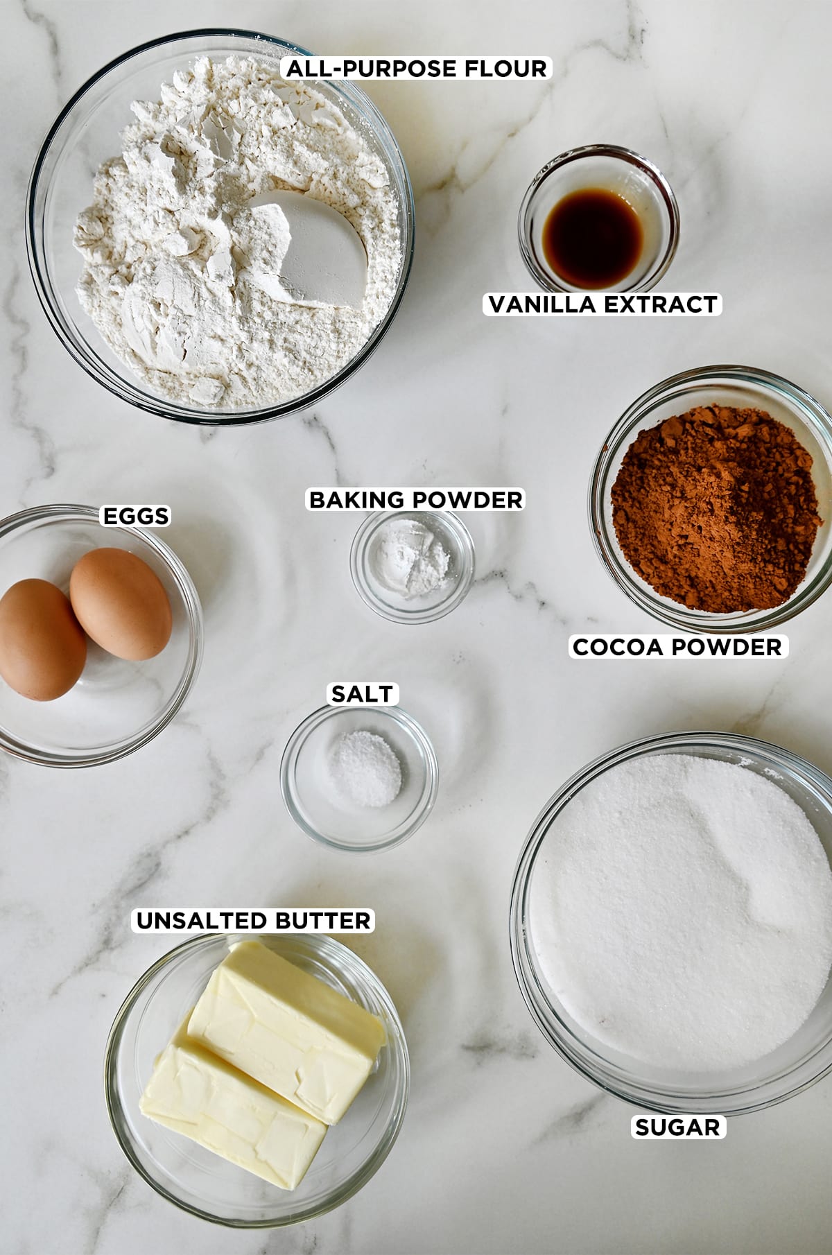 Various sizes of clear bowls containing ingredients to make chocolate sugar cookies, including vanilla extract, cocoa powder, sugar, butter, salt, baking powder, eggs and flour.
