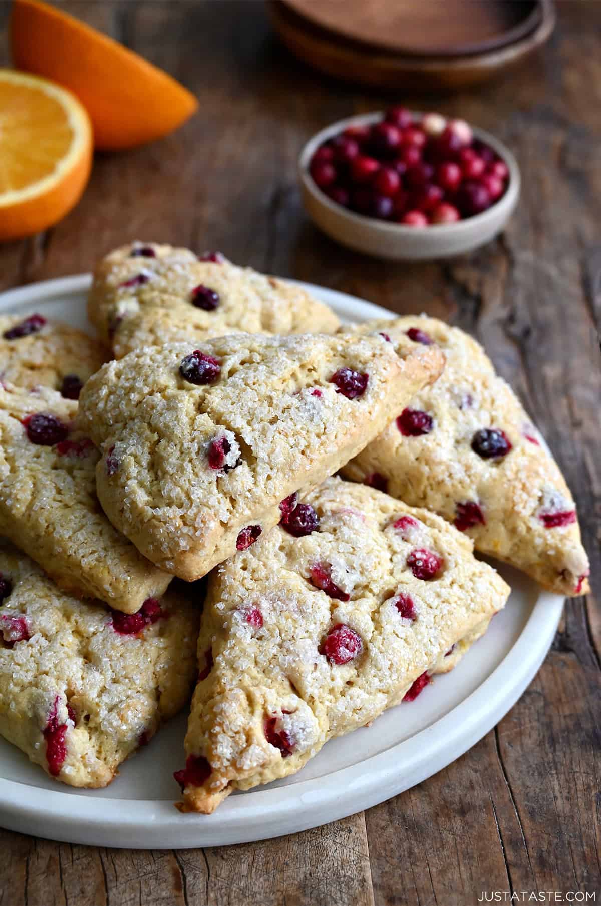 Cranberry orange scones with coarse sugar on a serving plate near a bowl of fresh cranberries and an orange that's been cut in half.