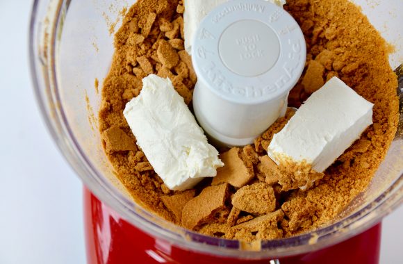 A food processor bowl containing crushed gingerbread cookies and cream cheese