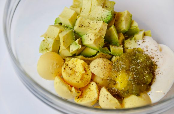 A clear bowl filled with cooked egg yolks, mustard, avocado, mayo and relish