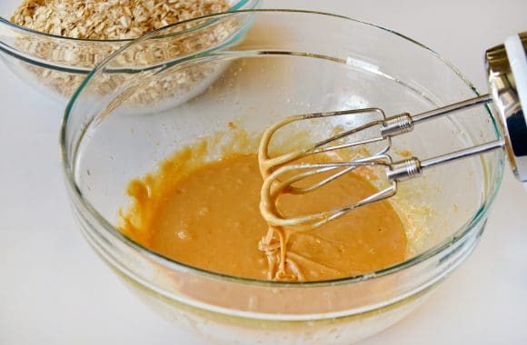 Large glass bowl with peanut butter, honey, egg, oil and vanilla extract with a handheld beater