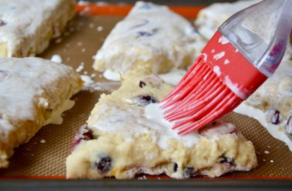 Orange cranberry scone on silpat lined baking sheet being brushed with heavy cream