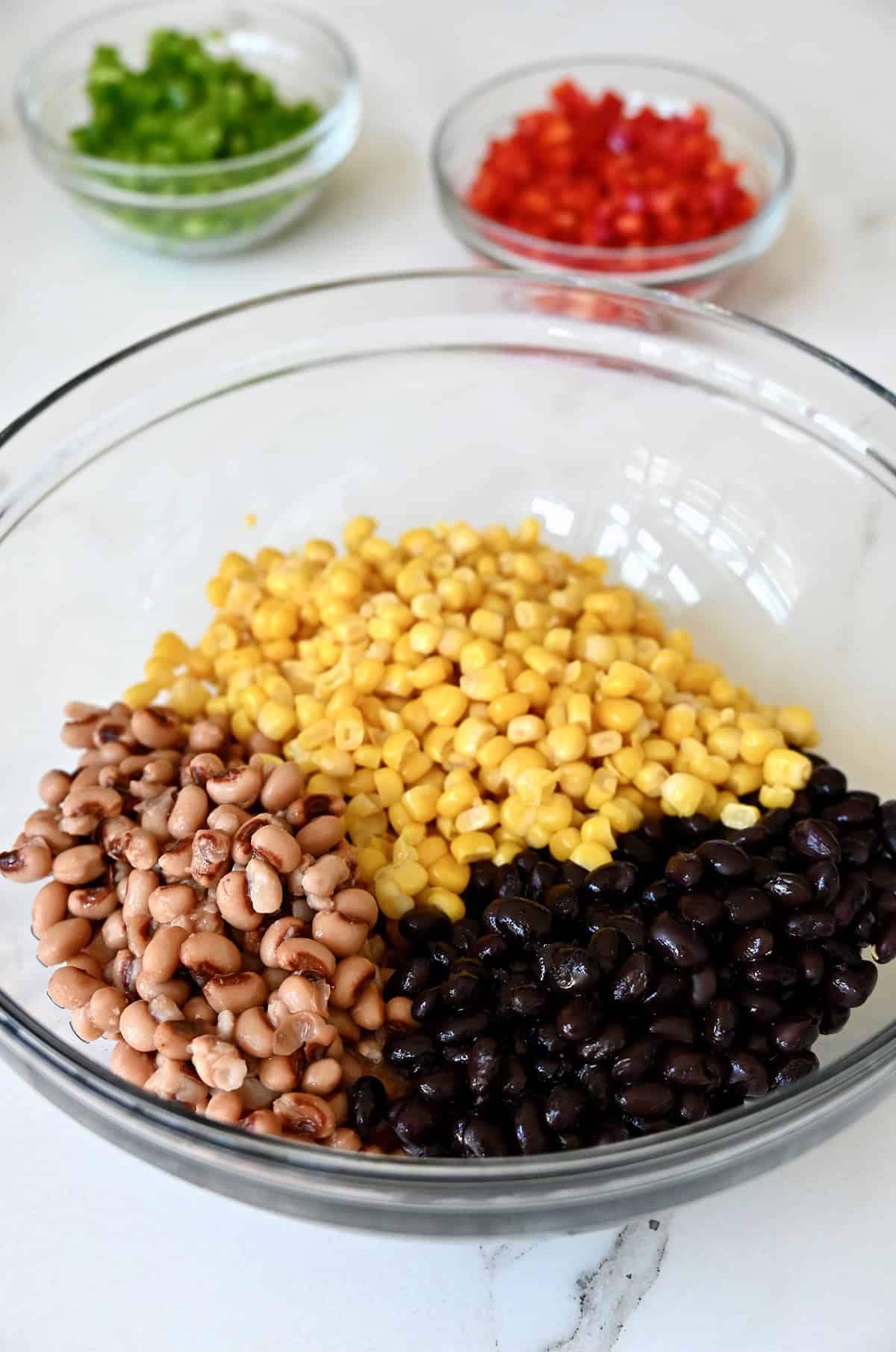 Black-eyed peas, corn and black beans in a large glass bowl with two small bowls containing diced red and green bell pepper nearby.