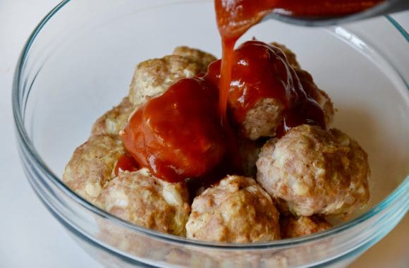 Baked meatballs in glass bowl with sweet and sour sauce being poured over them