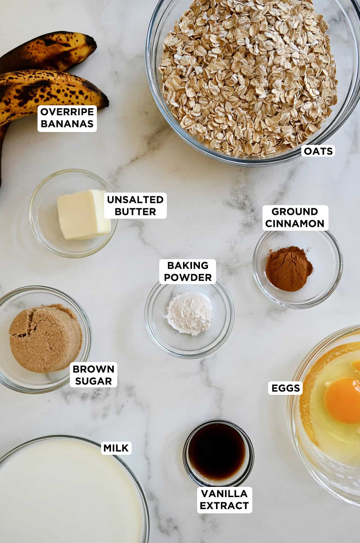 Ingredients to make baked oatmeal, including old-fashioned rolled oats, butter, brown sugar, eggs, overripe bananas, baking powder, milk and cinnamon.