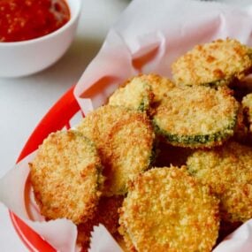 Baked Parmesan Zucchini Chips in red basket lined with white food paper with ketchup and mayonnaise in the background.